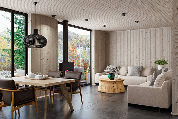 Warm and inviting Scandinavian-style living room captured from a new angle, showcasing two beige sofas arranged around a round solid-wood stump coffee table, decorated with minimalist candles for a cozy touch. The space features a freestanding modern black metal fireplace, a dining area with four chairs, and continuous pine wood paneling across the walls and ceiling. Matte black stone floor tiles add contrast and sophistication, enhancing the natural textures. Ideal for use in interior design promotions, eco-cabin marketing, Scandinavian lifestyle visuals, real estate, architectural presentations, and sustainable furniture or material branding.